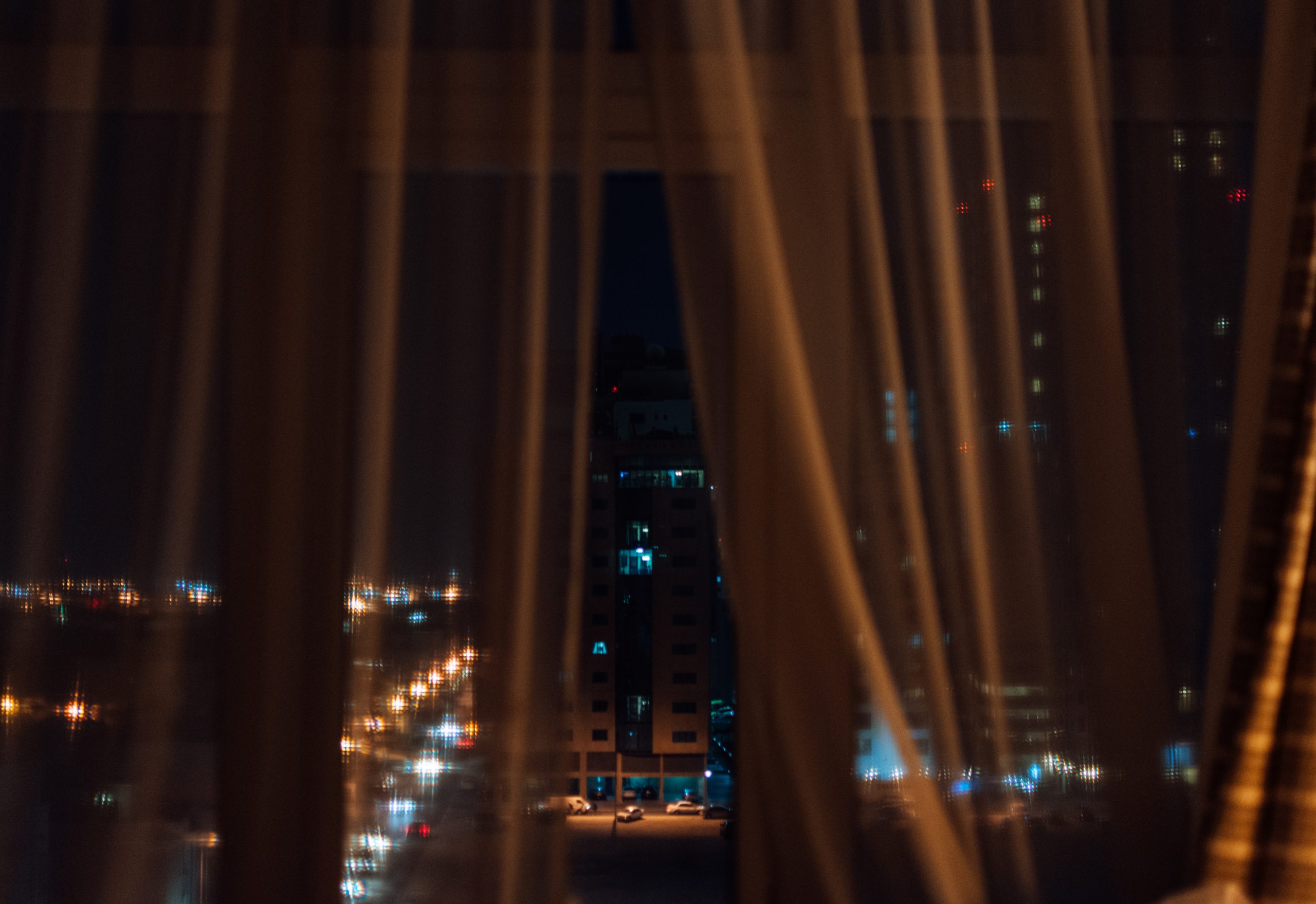 Photograph looking through a window encased by curtains, into a night urban scene.