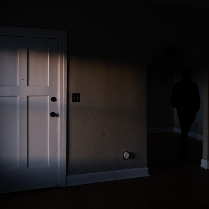 Photograph of a dark room in a house with a white door on the left and a dark figure in a doorway to the right